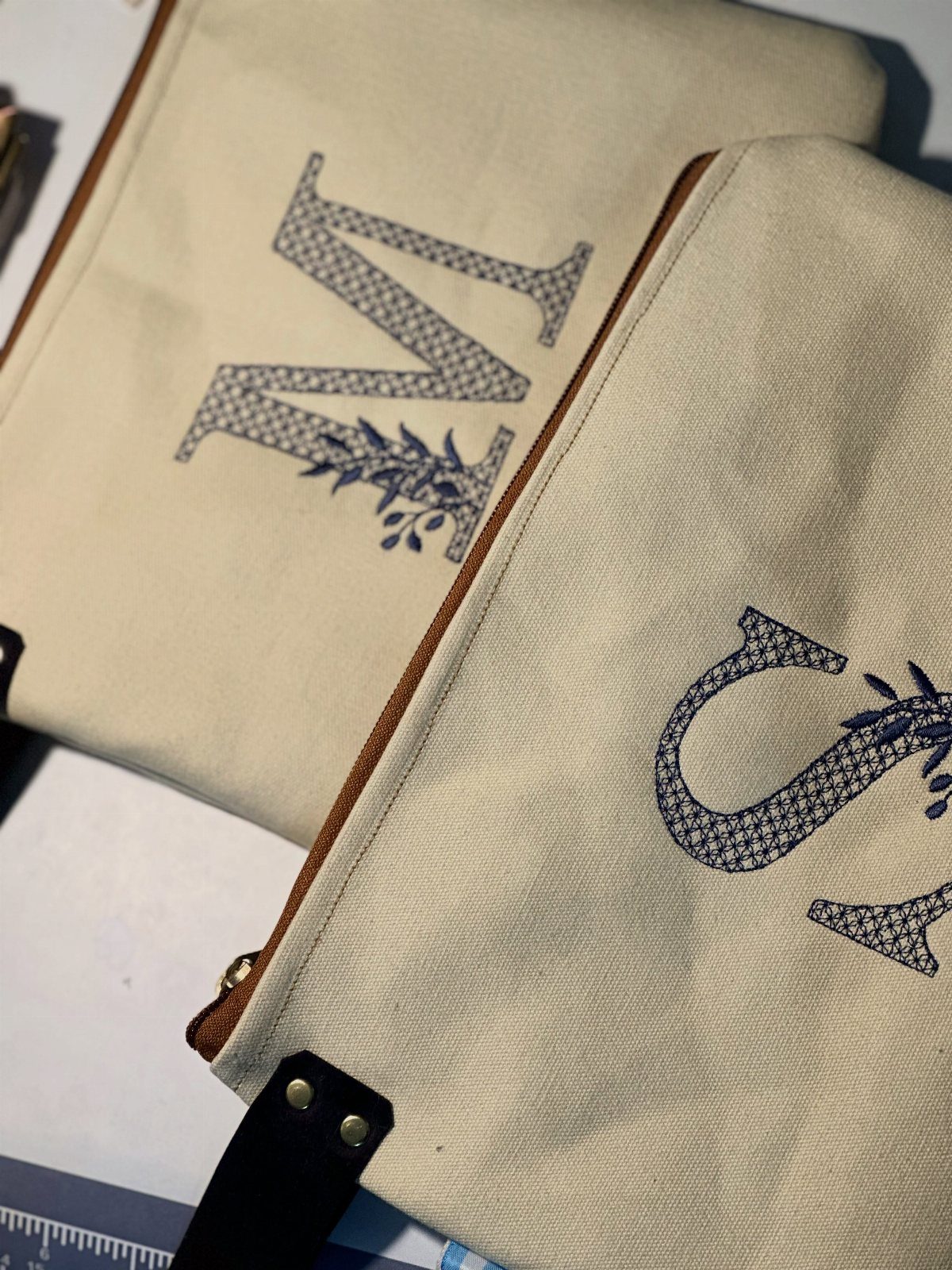 Two cream canvas pouches featuring blue patterned monogram letters with floral accents.