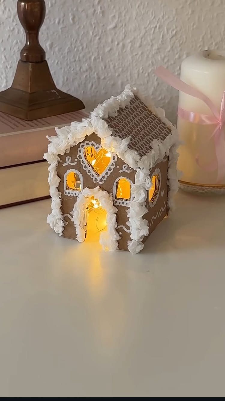 Handmade cardboard gingerbread house with white trim and warm glowing fairy lights inside.