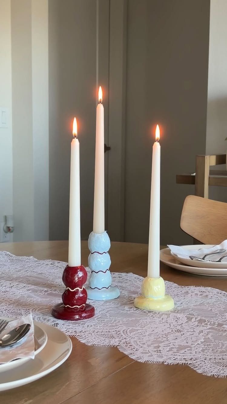 Three lit white taper candles in red, blue, and yellow ceramic holders on lace.