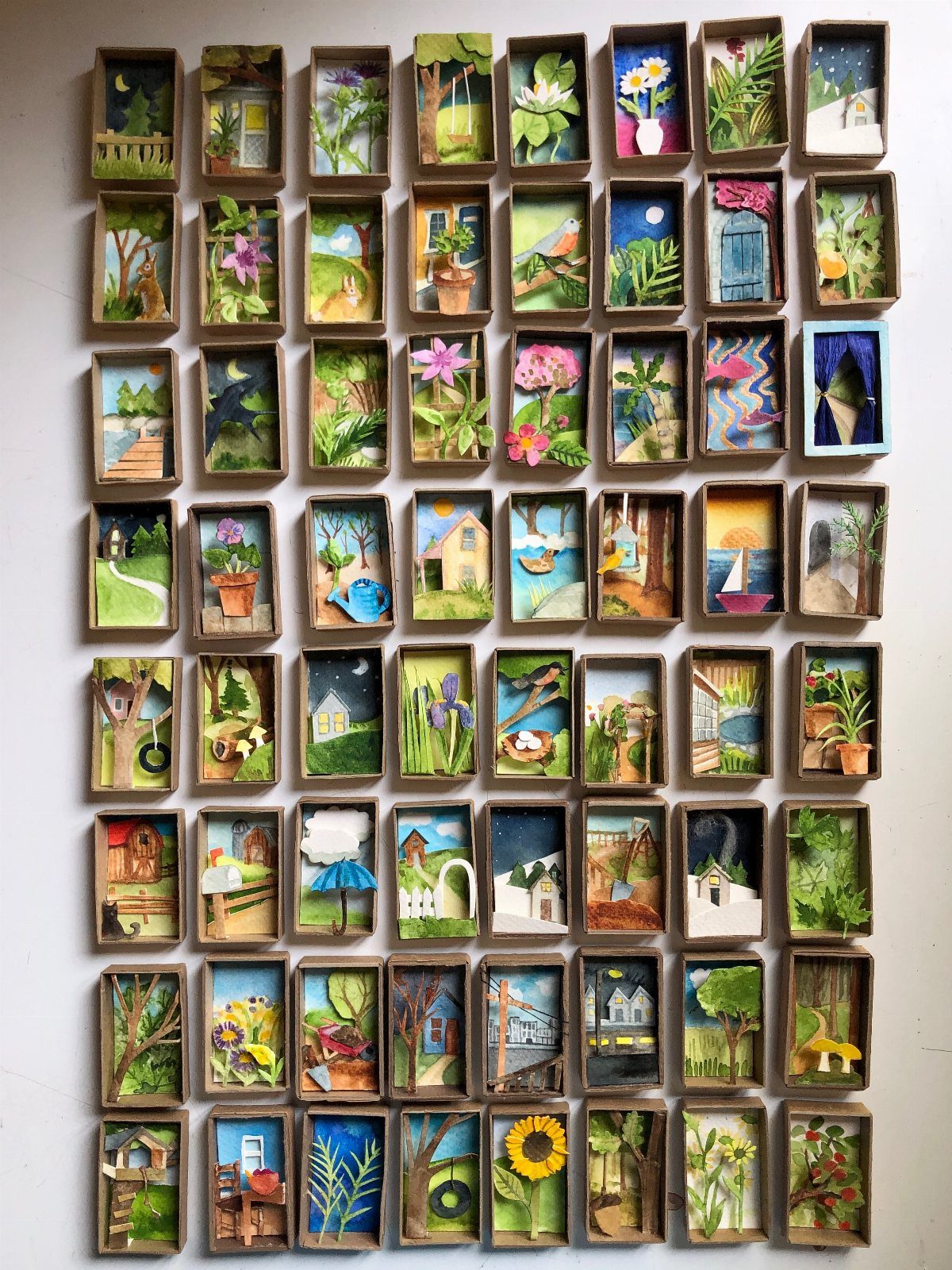 Grid of miniature 3D landscape scenes inside small cardboard boxes on a wall