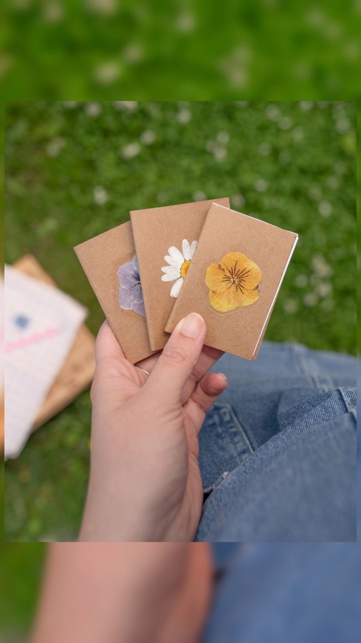 Three small brown kraft paper notebooks decorated with pressed flowers held in a hand.