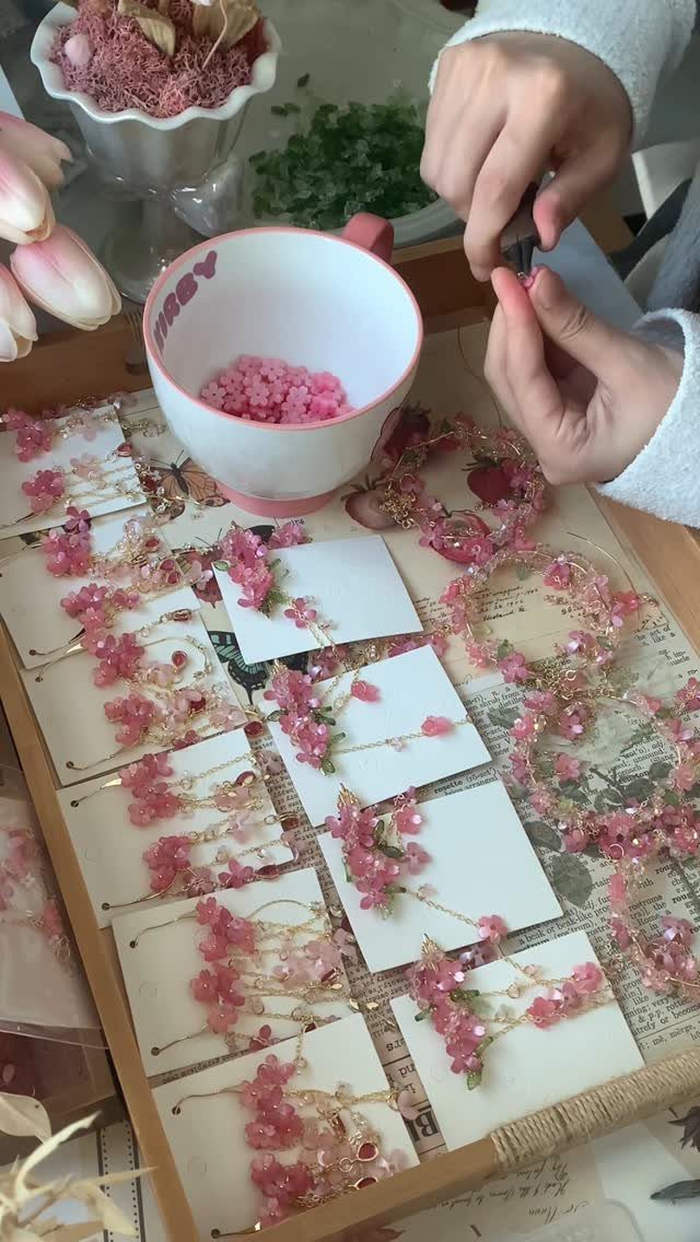 Hands assembling pink beaded necklaces with floral accents on a wooden tray.