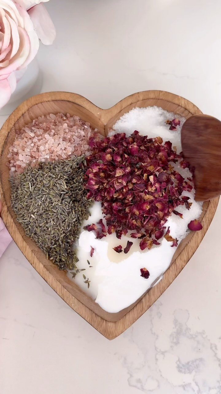 Heart-shaped wooden bowl filled with pink and white salts, herbs, and rose petals.