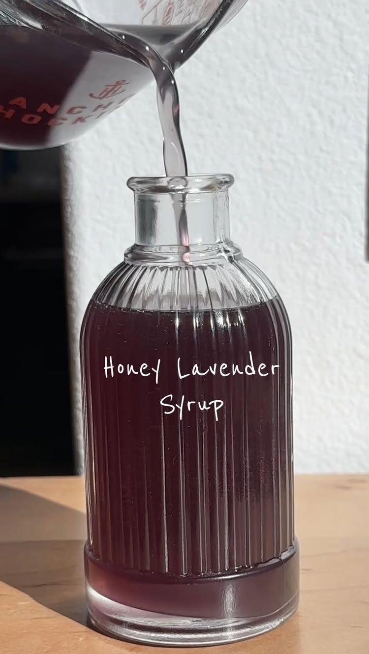 Dark purple honey lavender syrup pouring into a ribbed glass bottle on wood.