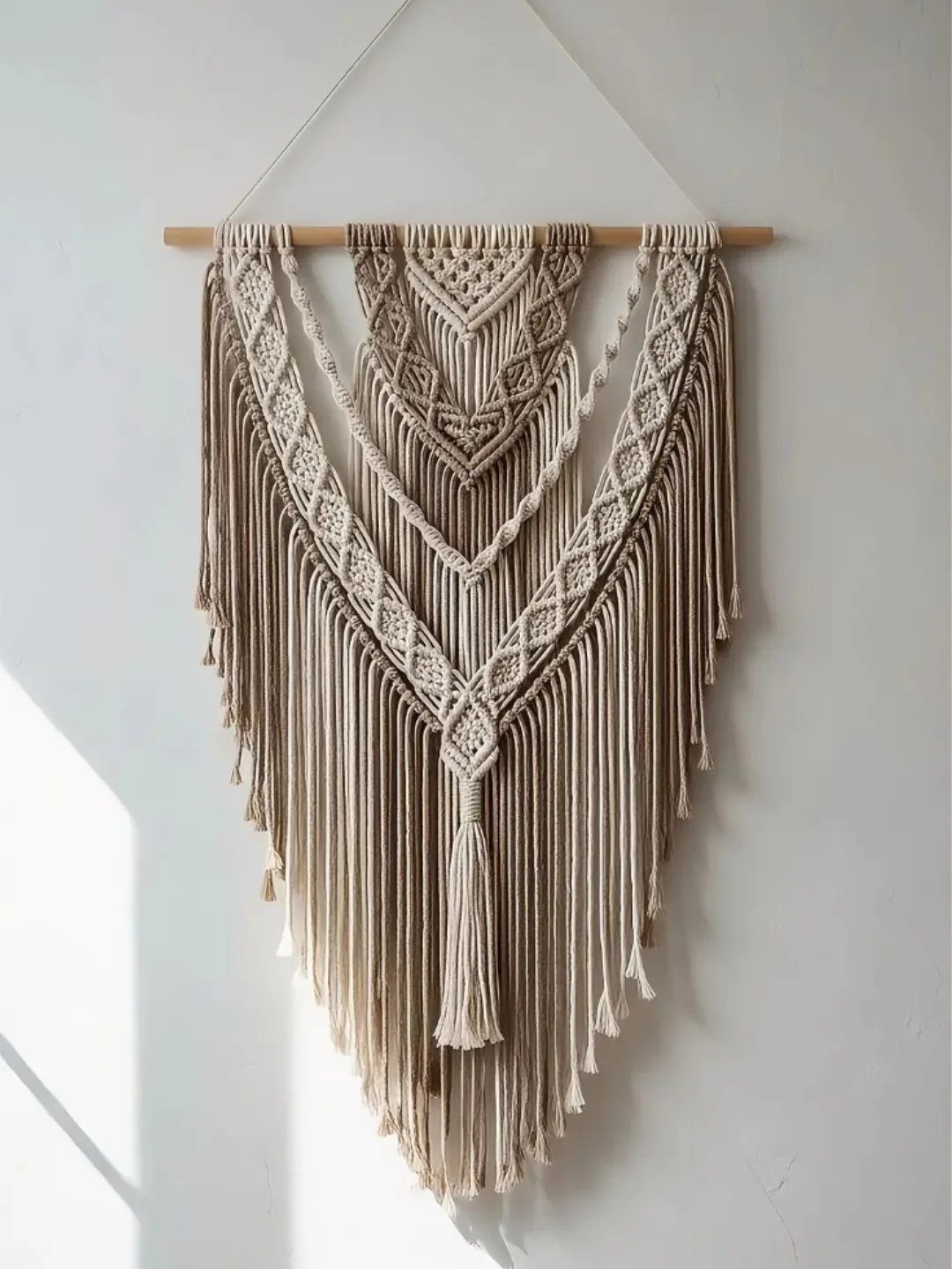 Beige macramé wall hanging with intricate knots and fringes on a white wall