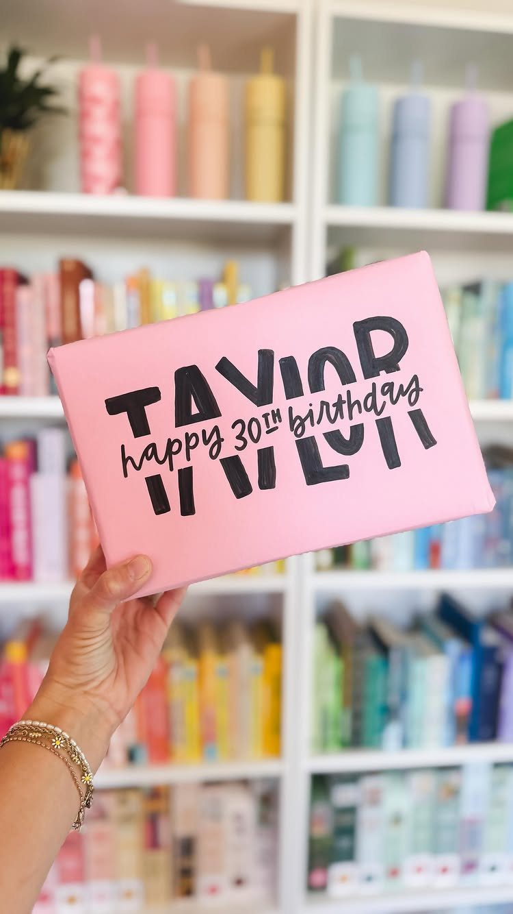 Pink wrapped gift with hand-lettered birthday message held against a colorful bookshelf background