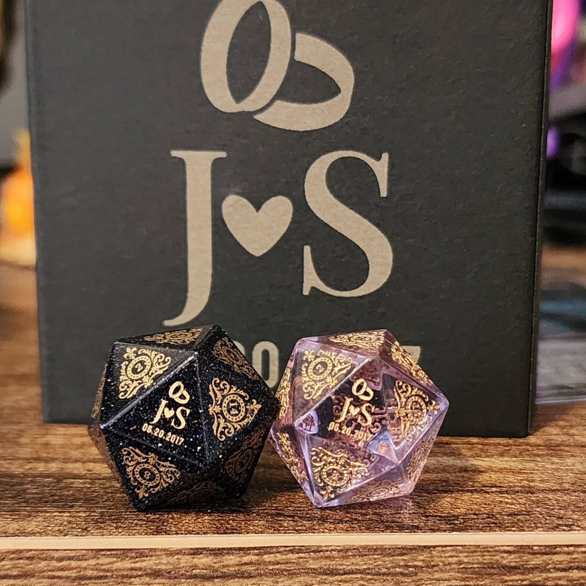 Two glittery twenty-sided dice with personalized initials and dates on a wooden surface.
