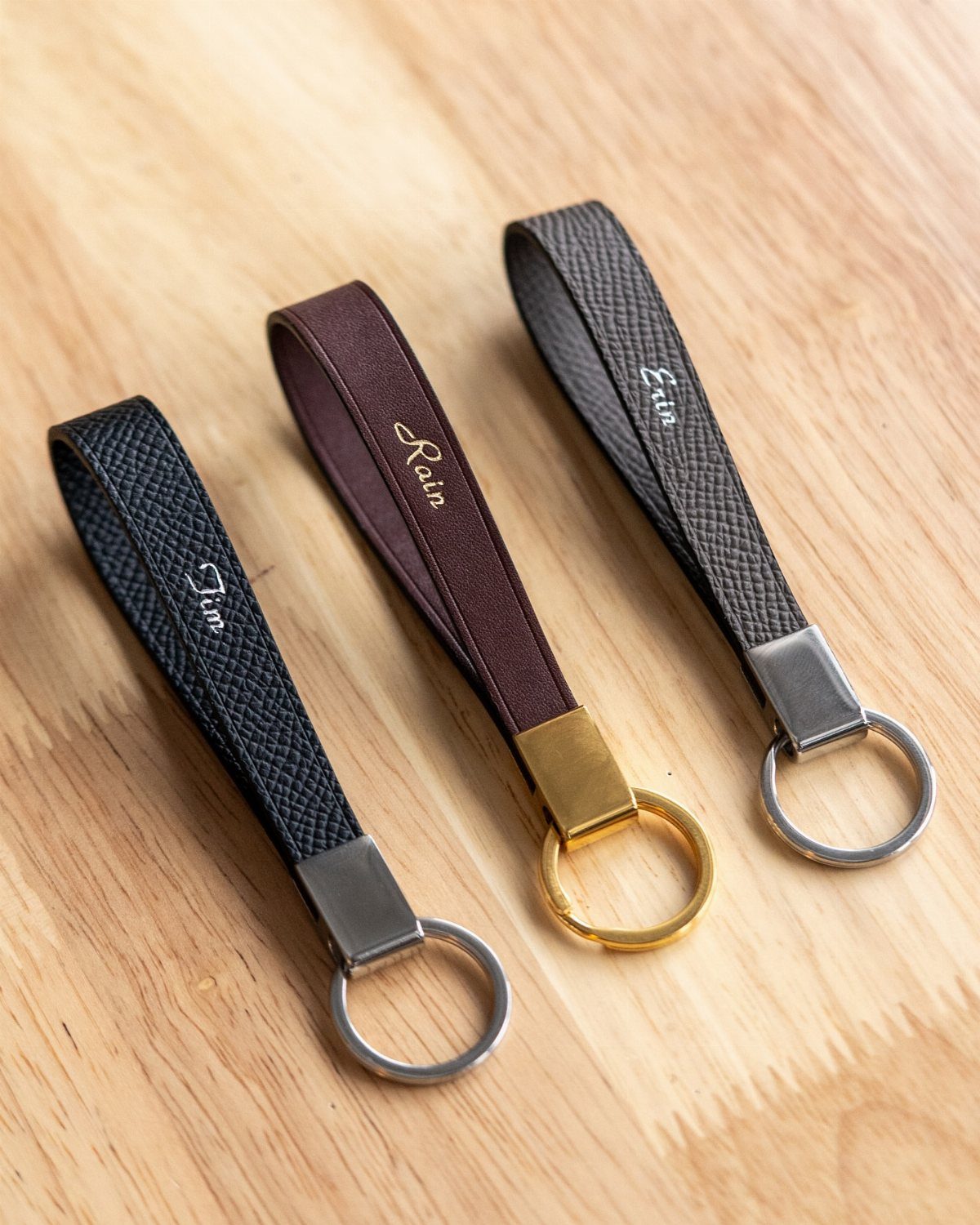 Three leather keychains with engraved names resting on a light wooden background.