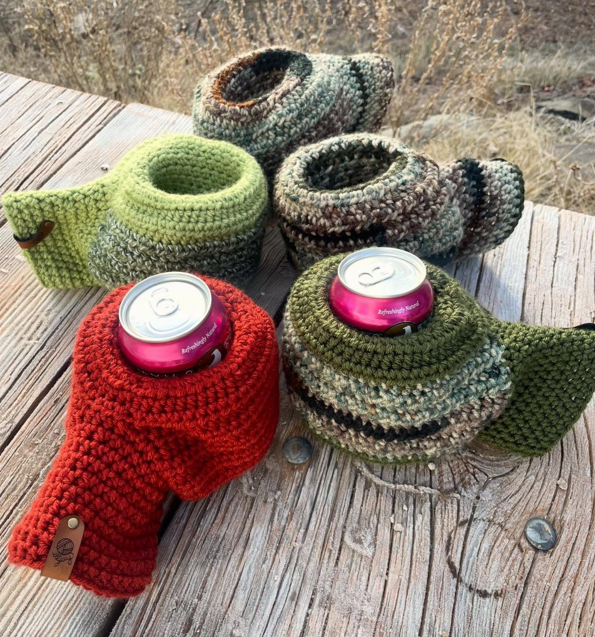 Crocheted beverage cosies in red and green holding aluminum cans on wooden table