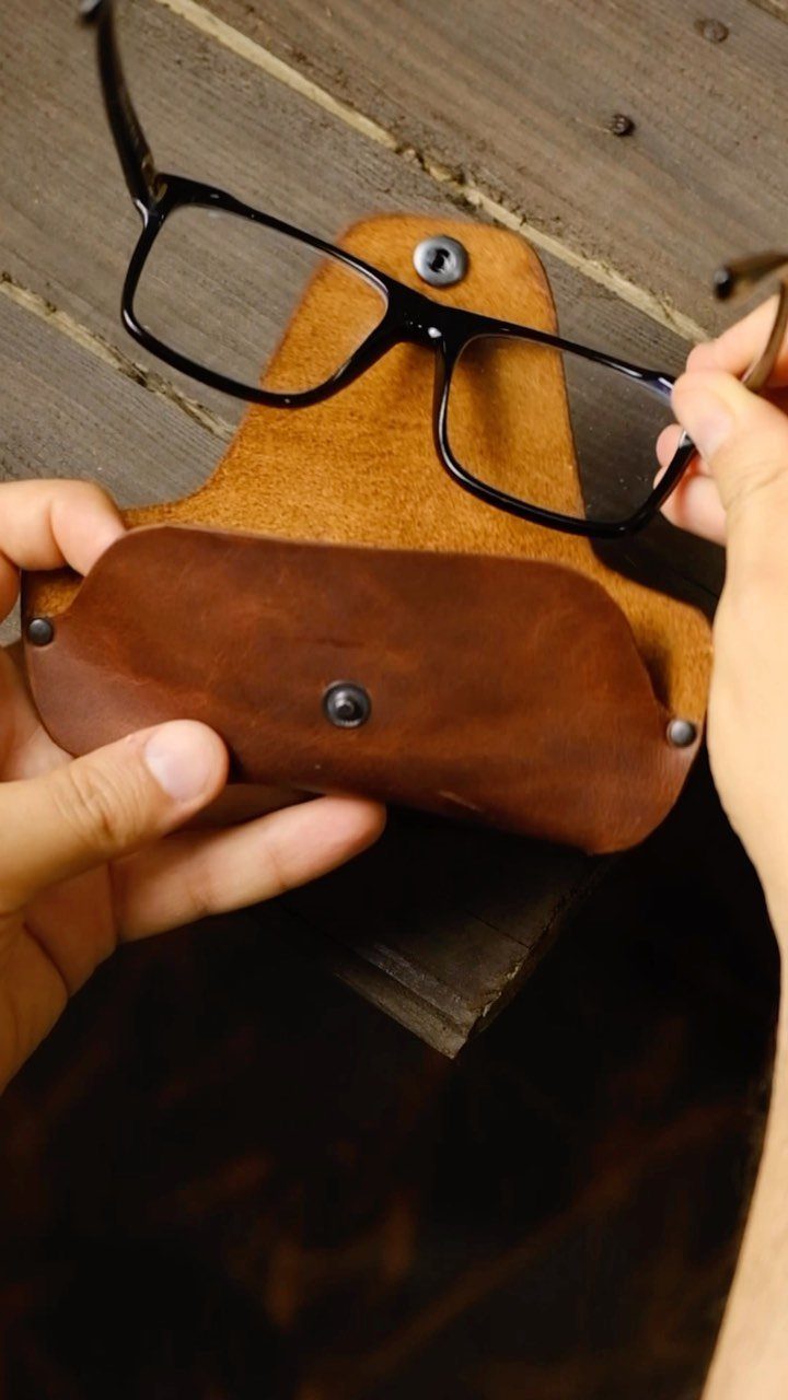 Brown leather glasses case and black-rimmed glasses held over a wooden background.