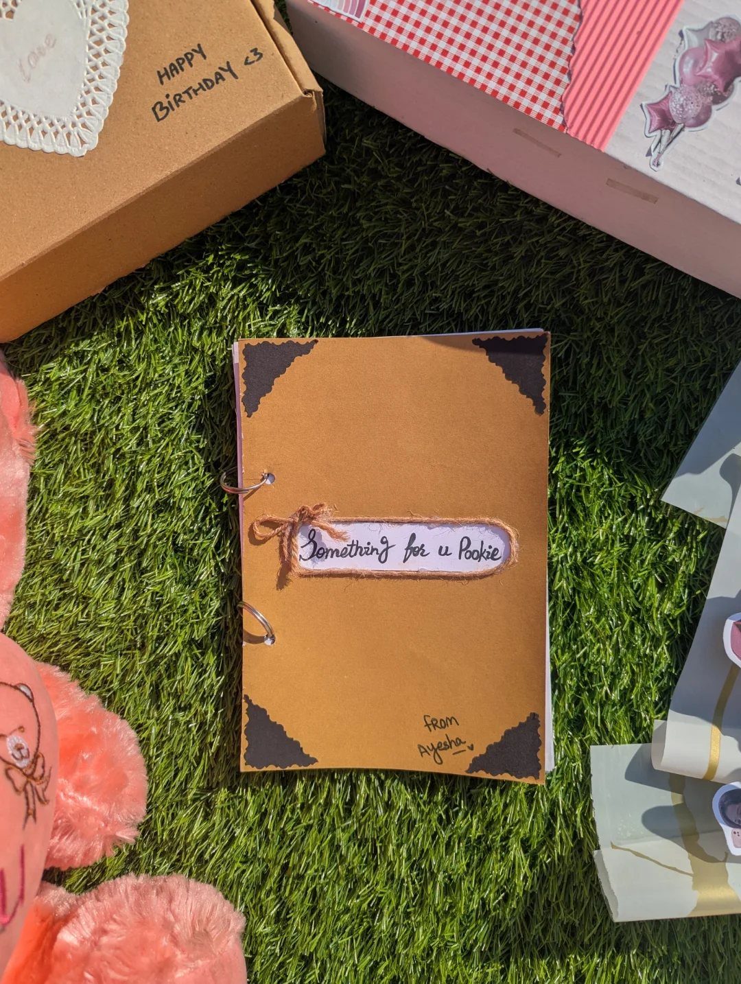 Handmade brown scrapbook with a personalized label resting on green grass near gift boxes