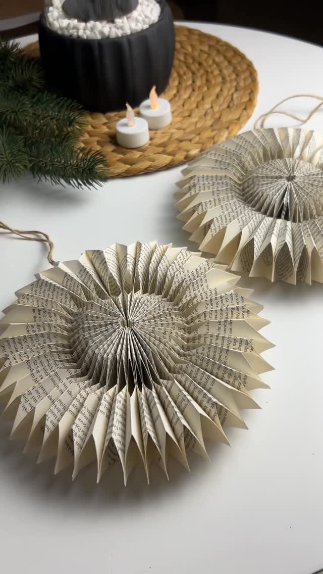 Two pleated book page ornaments on a white table with candles and pine.