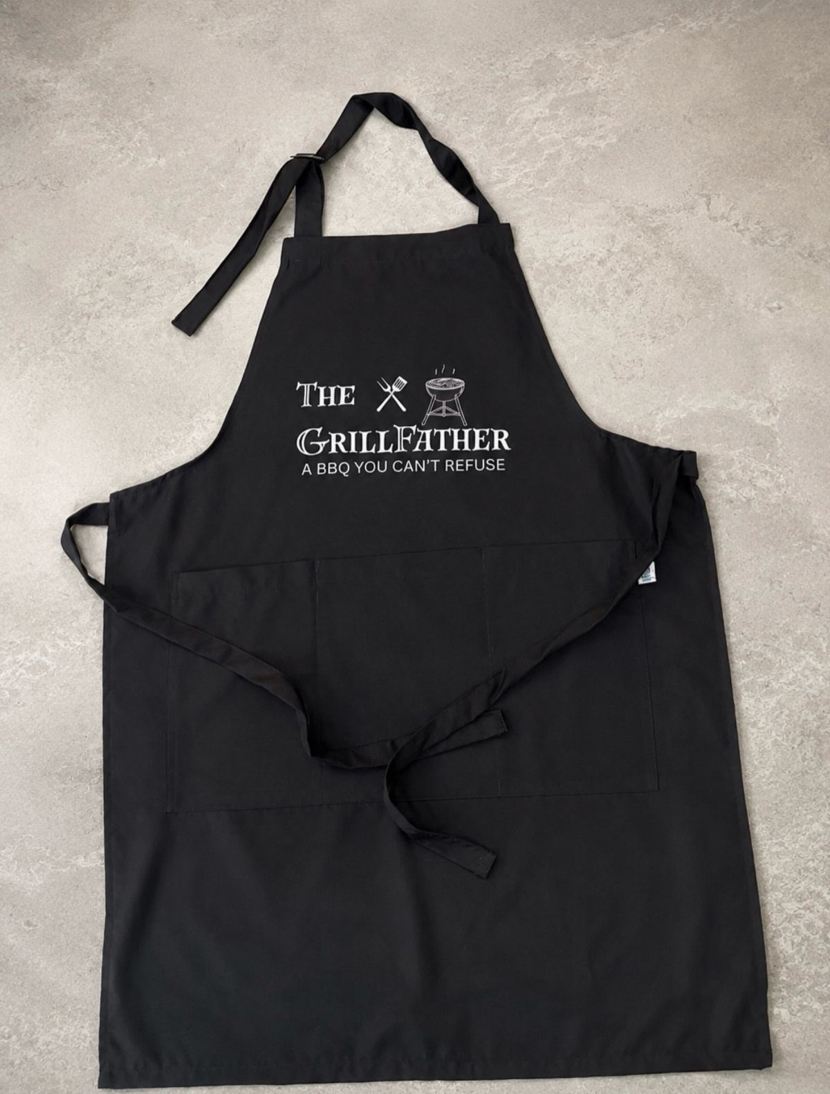 Black apron with white Grillfather text and BBQ icons on grey background