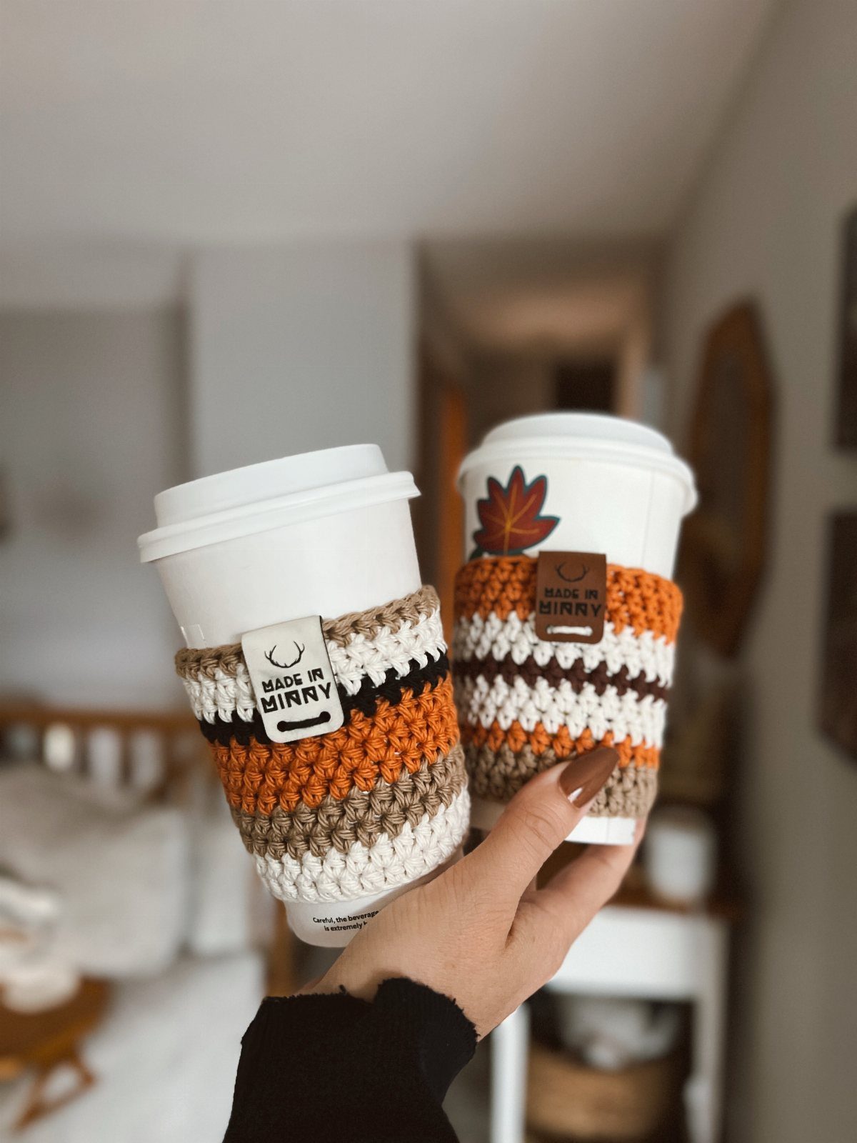 Two white coffee cups with orange and brown crocheted sleeves held by a hand.