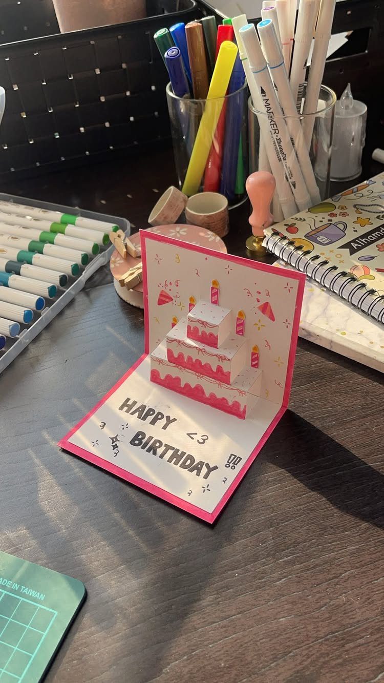 Pop-up birthday card with a pink tiered cake on a wooden desk.