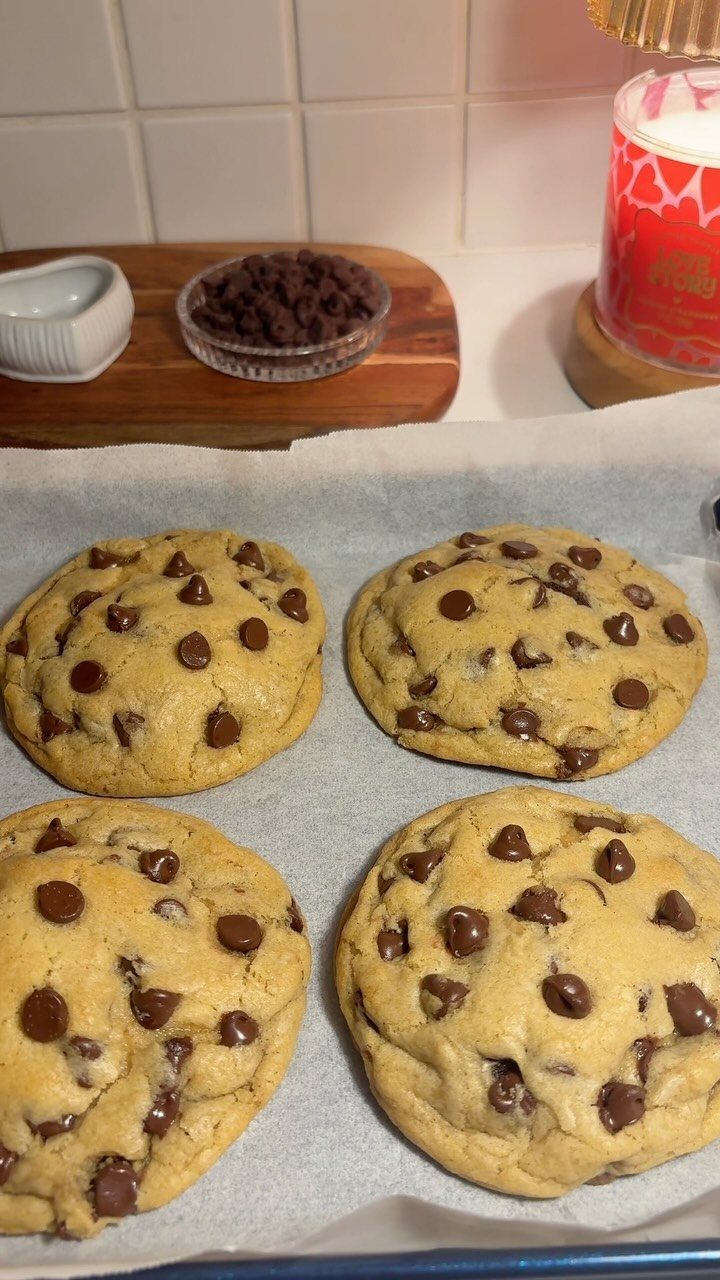 Four chocolate chip cookies on parchment paper with chocolate chips in background