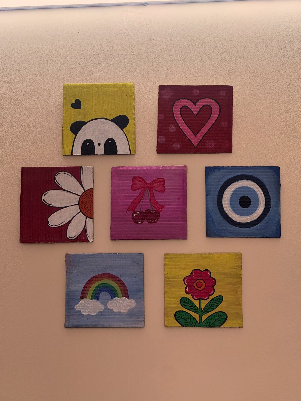 Eight small square canvases with colorful painted illustrations hanging on a white wall.