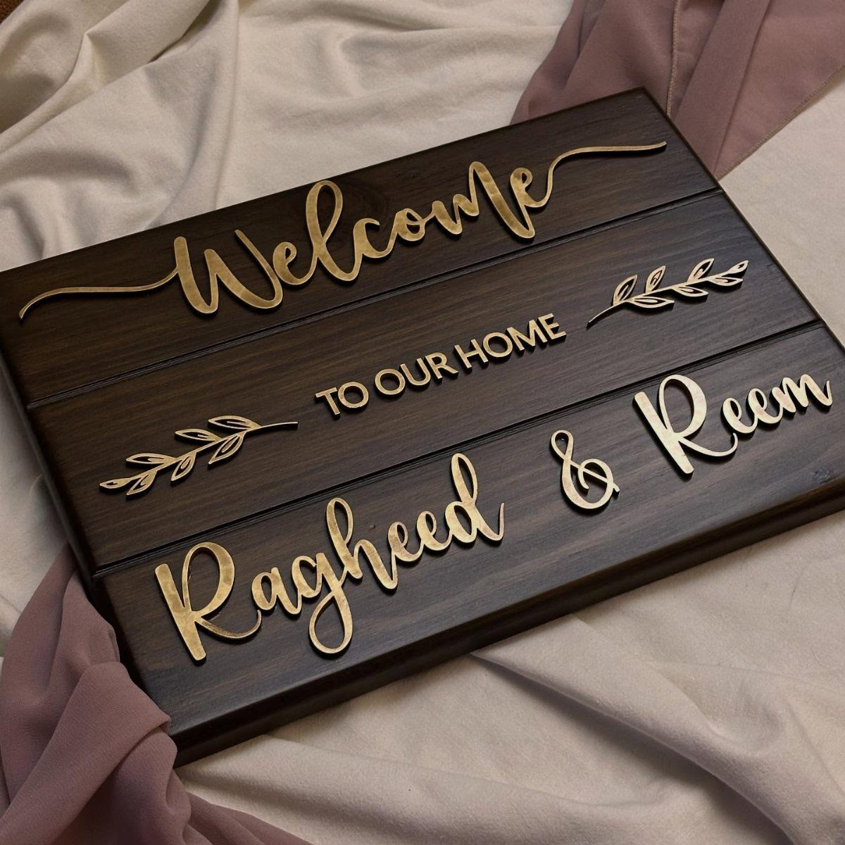 Wooden welcome sign with gold lettering resting on soft white and mauve fabric.