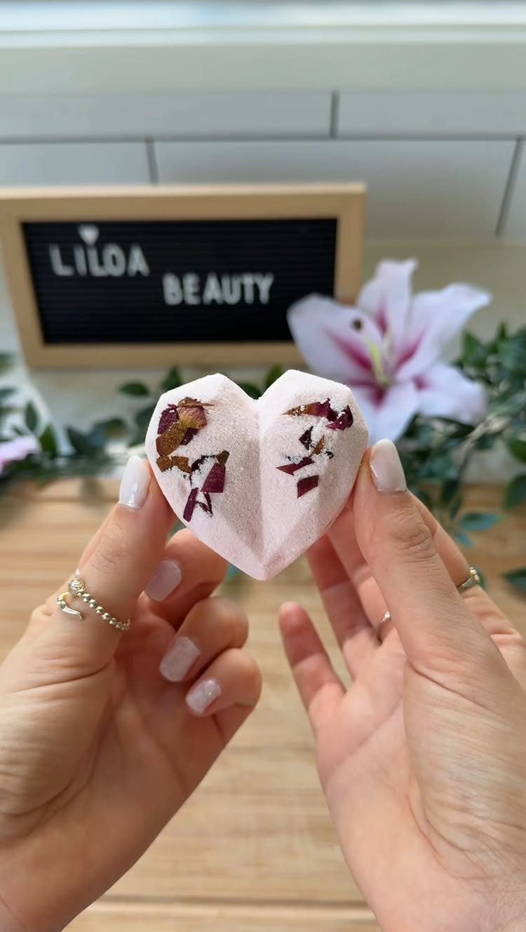 Hands holding a pink heart-shaped bath bomb with dried flower petals on top.