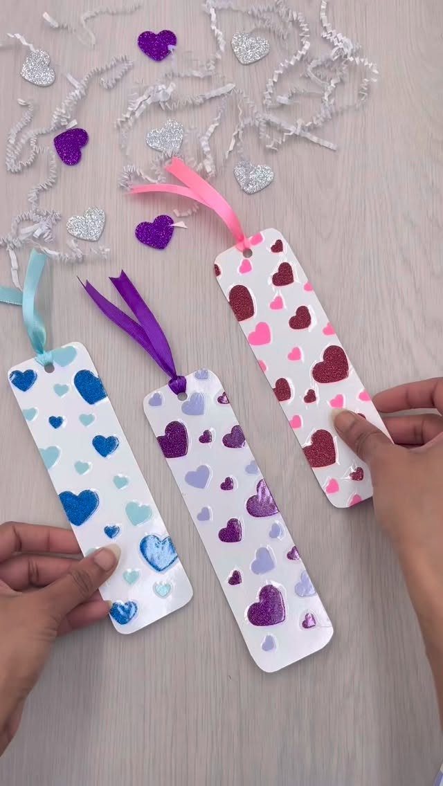 Three white bookmarks with colorful glitter hearts and ribbons held by two hands.