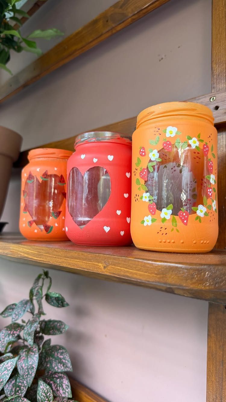 Three painted red and orange jars with heart cut-outs on a wooden shelf.