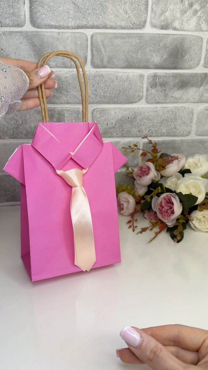 Pink paper gift bag shaped like a shirt with a satin tie and twine handles.