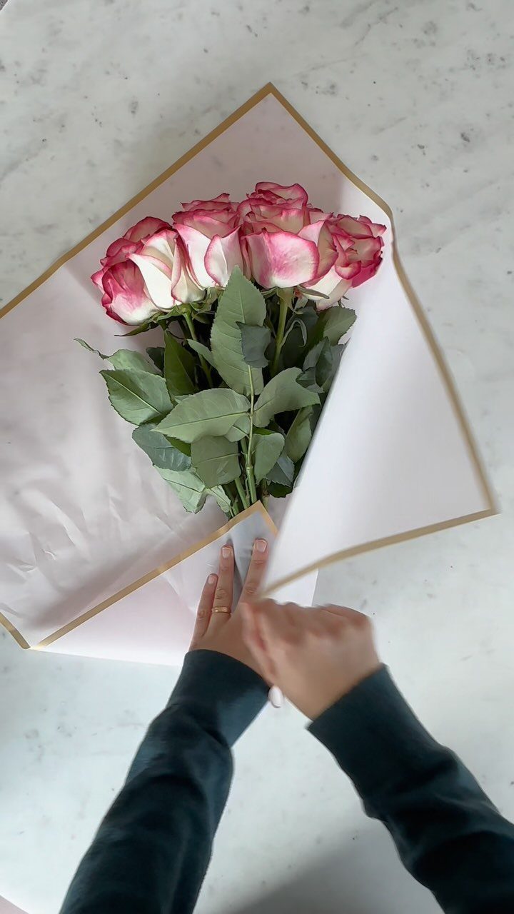 Pink and white roses wrapped in gold-edged pink paper held by two hands.