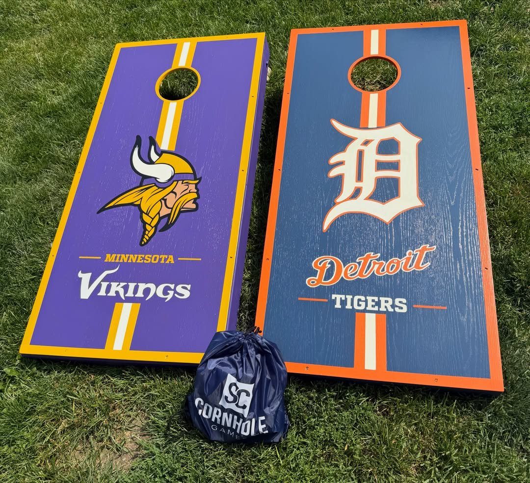 Two sports-themed corn hole boards and a bean bag pouch on green grass.