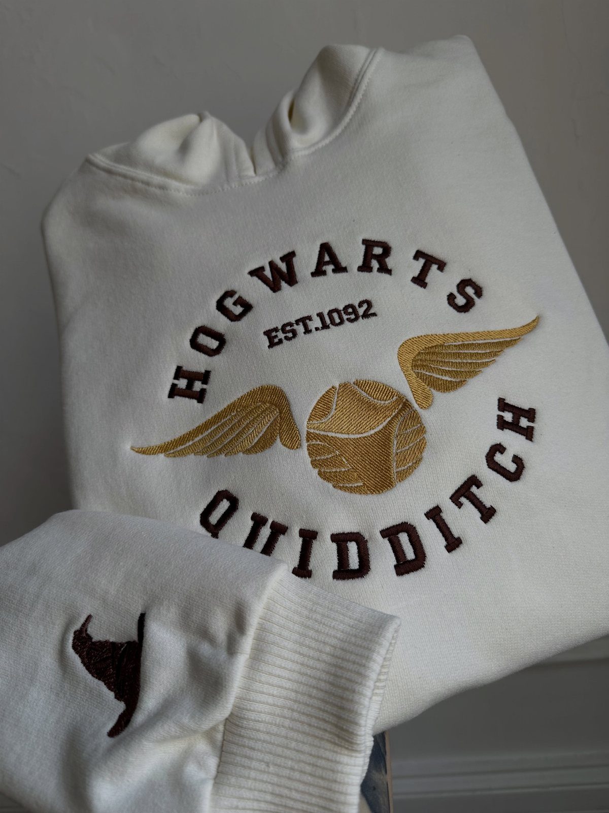White crewneck sweatshirt with brown Hogwarts Quidditch embroidery and a golden winged ball.