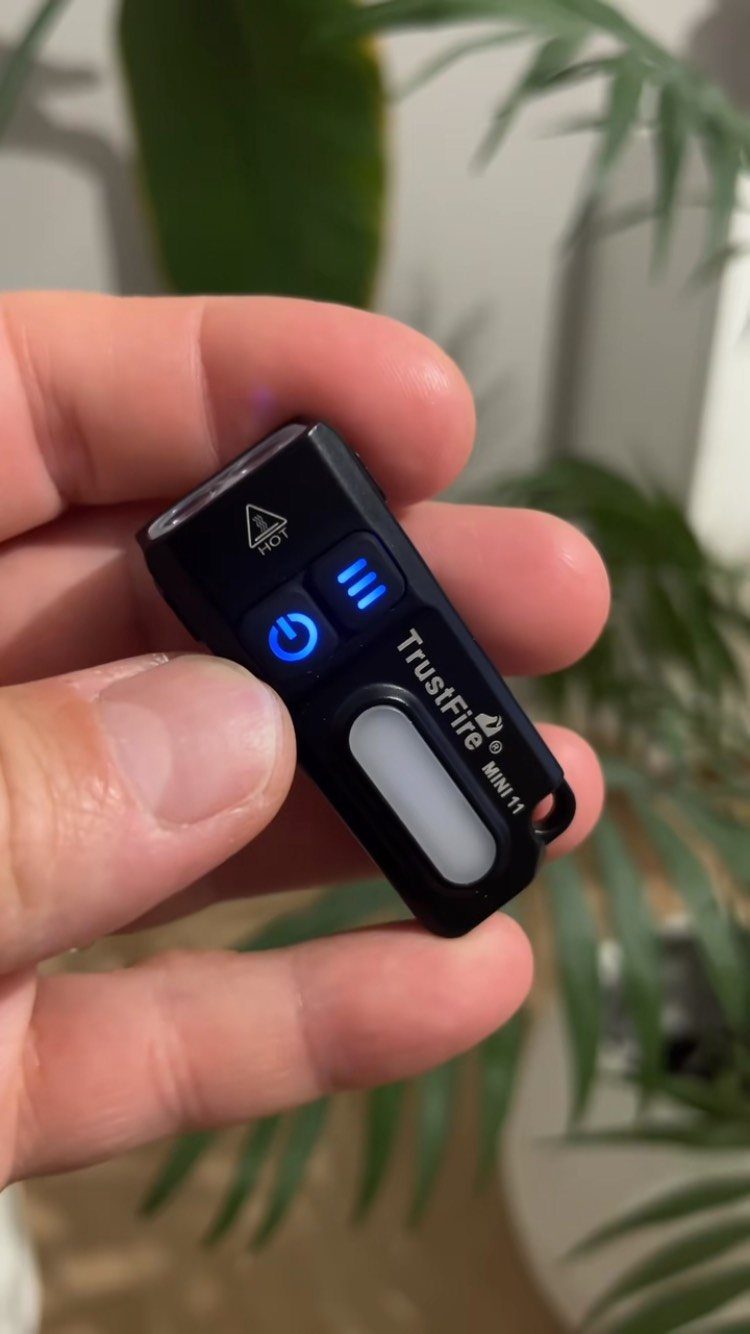 Small black TrustFire mini flashlight held between fingers with a glowing blue button.