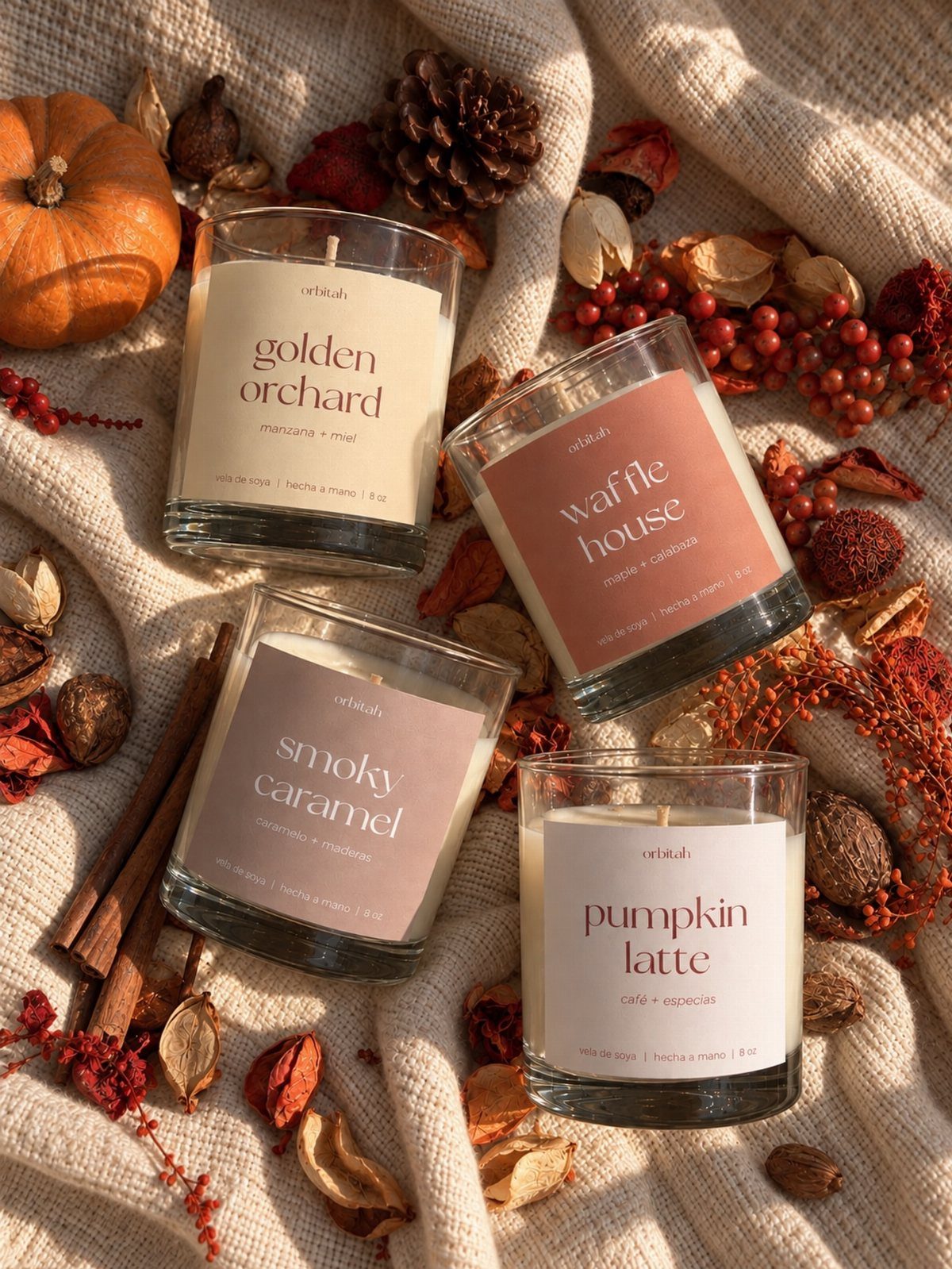 Four scented candles in glass jars surrounded by autumn leaves and a pumpkin.