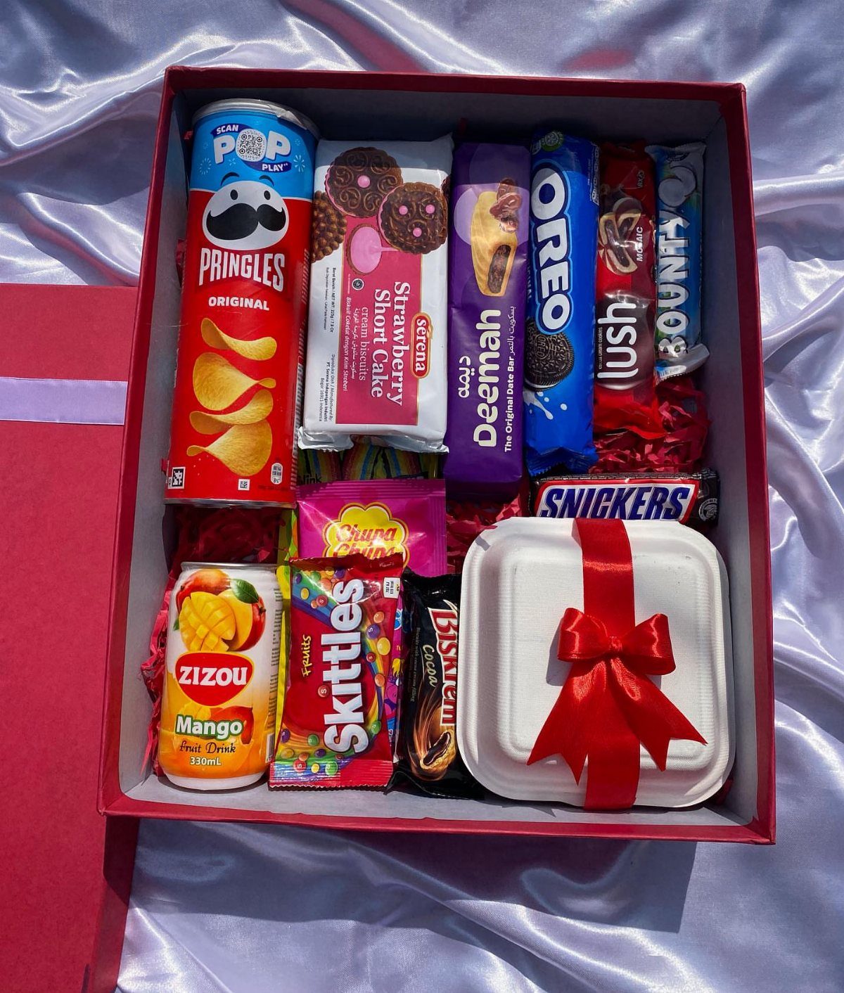 Red gift box filled with assorted snacks and a ribboned white container.