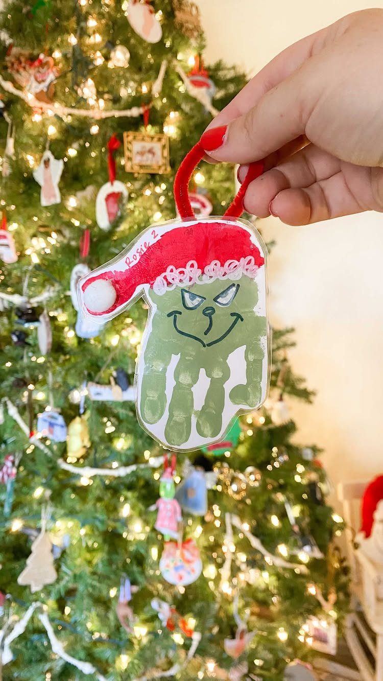 Handprint ornament painted as the Grinch held in front of a Christmas tree