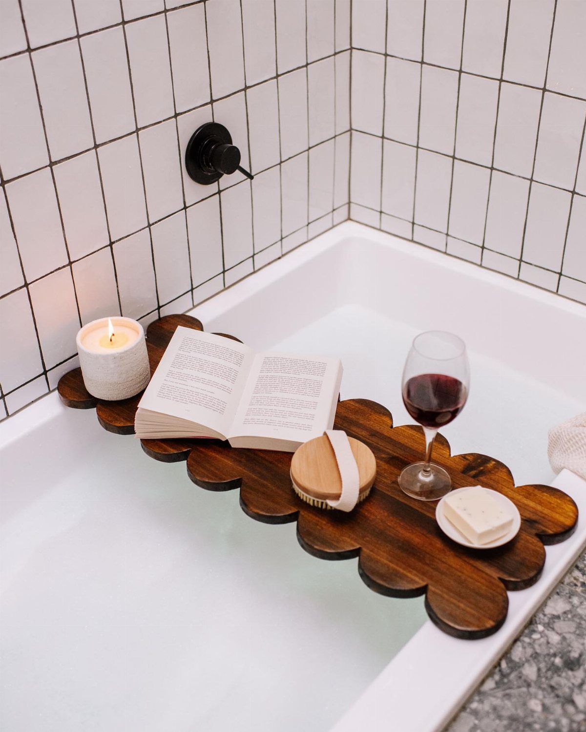 Wooden scalloped bath tray with a candle, book, wine, and soap across a tub