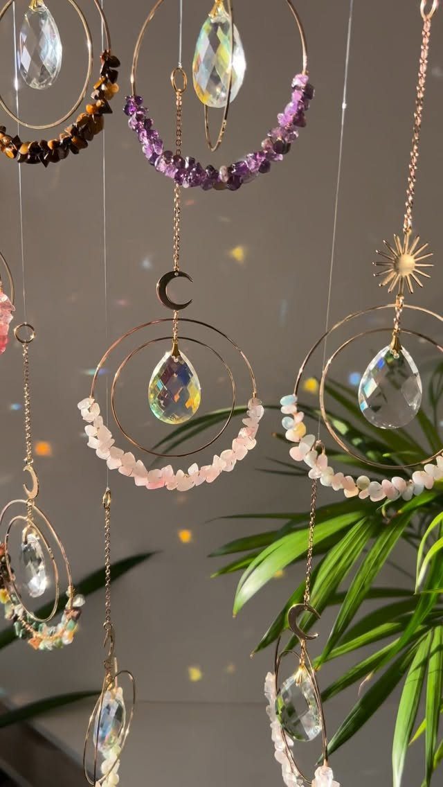 Hanging suncatchers with gold rings, iridescent crystals, and colorful gemstones near green leaves.