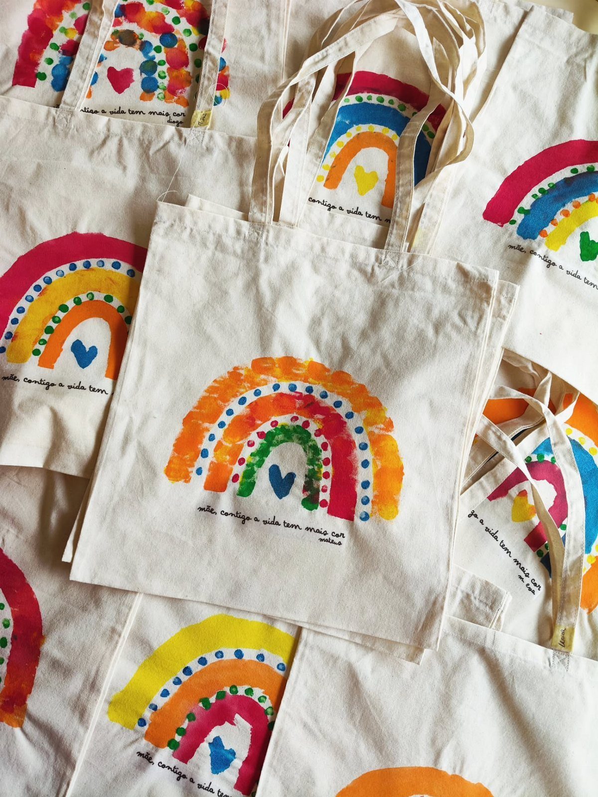 White canvas tote bags with colorful hand-painted rainbows and small blue hearts.
