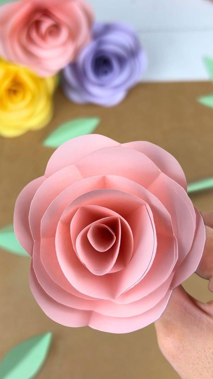 Pink cardstock paper rose held by a hand with other colorful paper flowers.