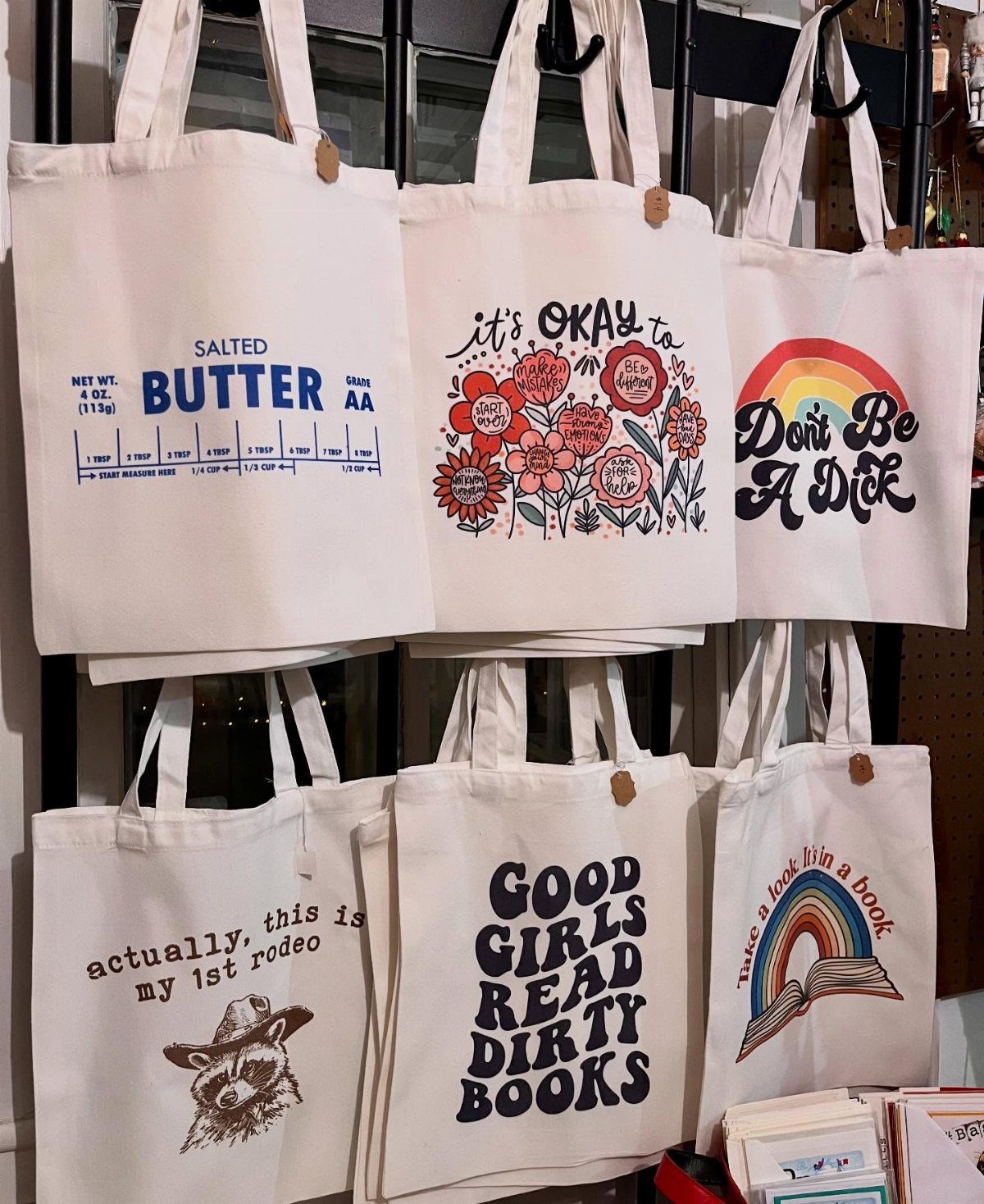 White canvas tote bags with various colorful graphic prints hanging on a display rack.