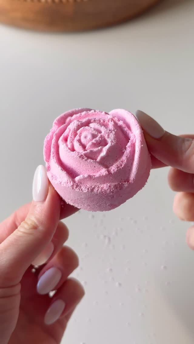 Pink rose-shaped bath bomb held by two hands against a white background.