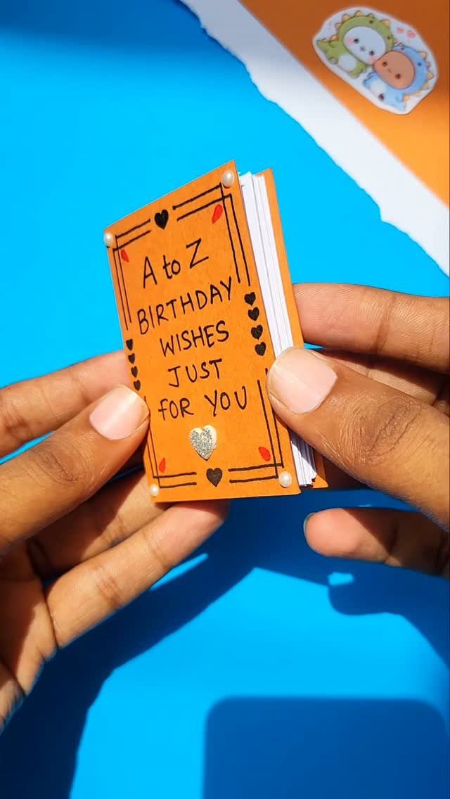 Handmade orange birthday wishes notebook held by two hands against a blue background.