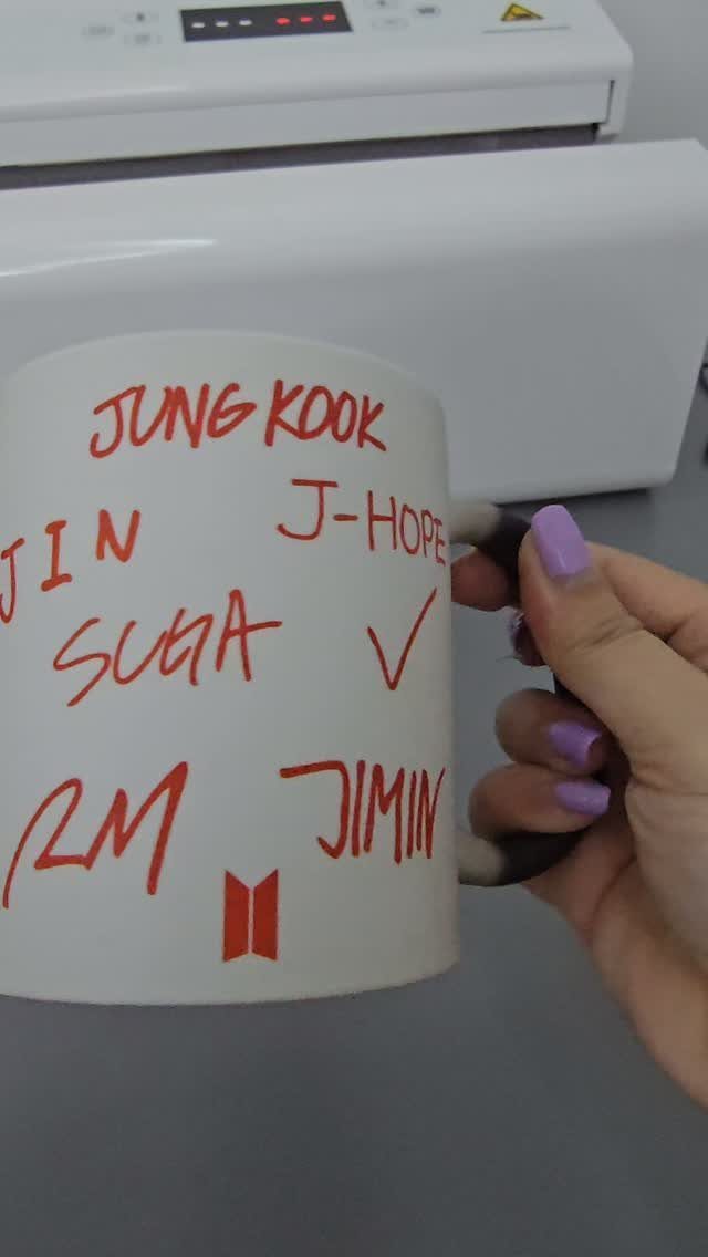White ceramic mug with red handwritten names held by a hand with purple nails.