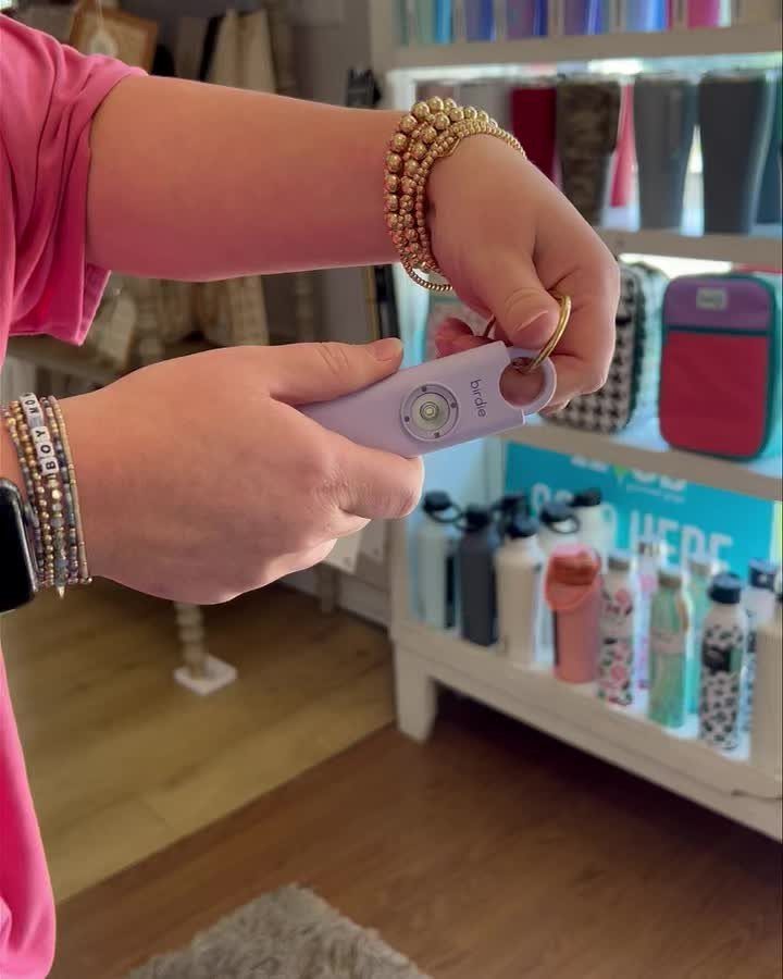 Hands holding a light purple personal safety alarm keychain with gold bracelets.
