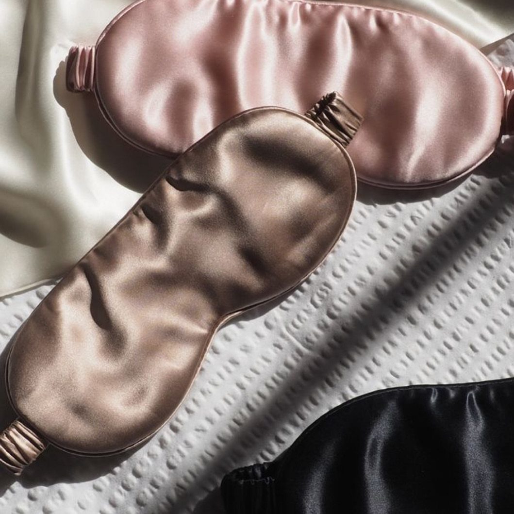 Three satin sleep masks in pink, champagne, and black on white textured fabric.