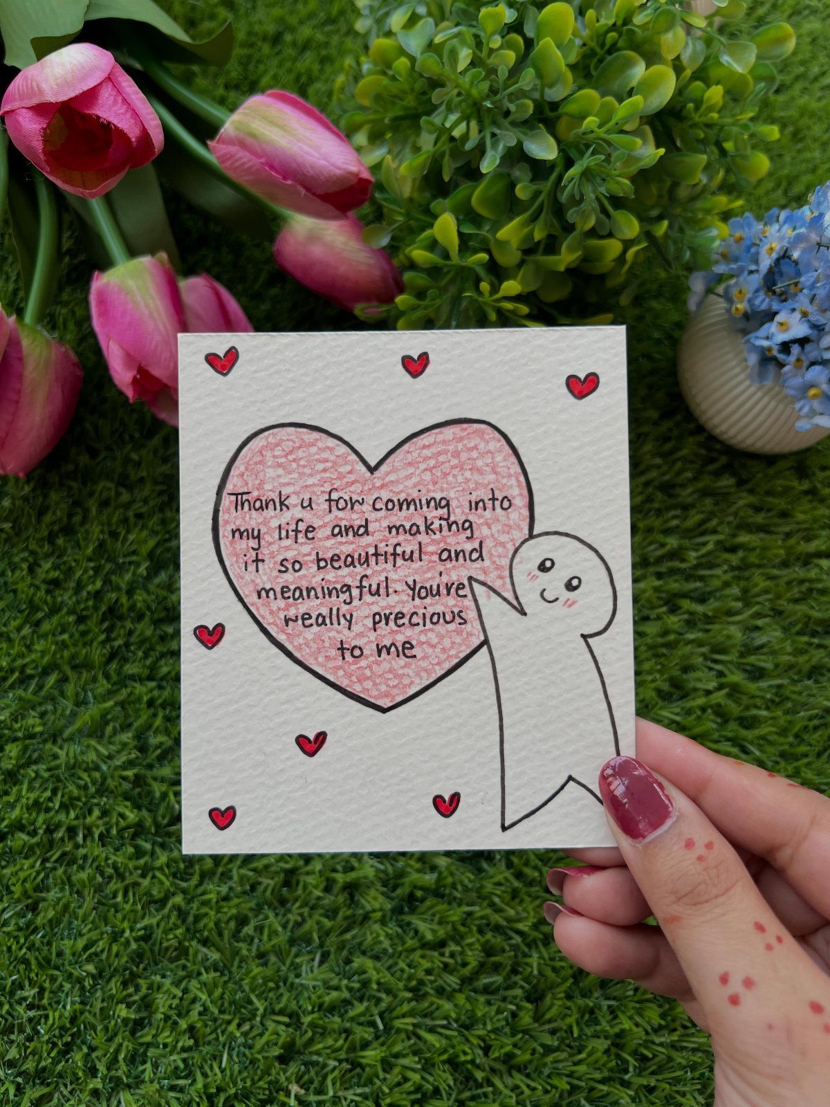 Handwritten card with a pink heart and cute character held over green grass.