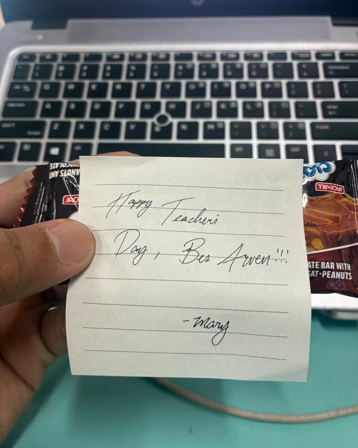Handwritten note for Teacher's Day held over a chocolate bar near a laptop.