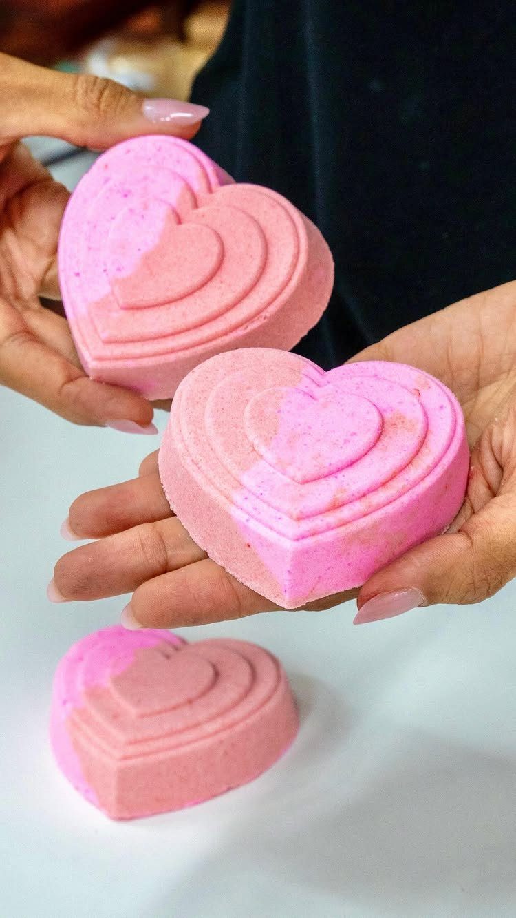 Pink heart-shaped bath bombs with concentric ridges held in a person's hands.