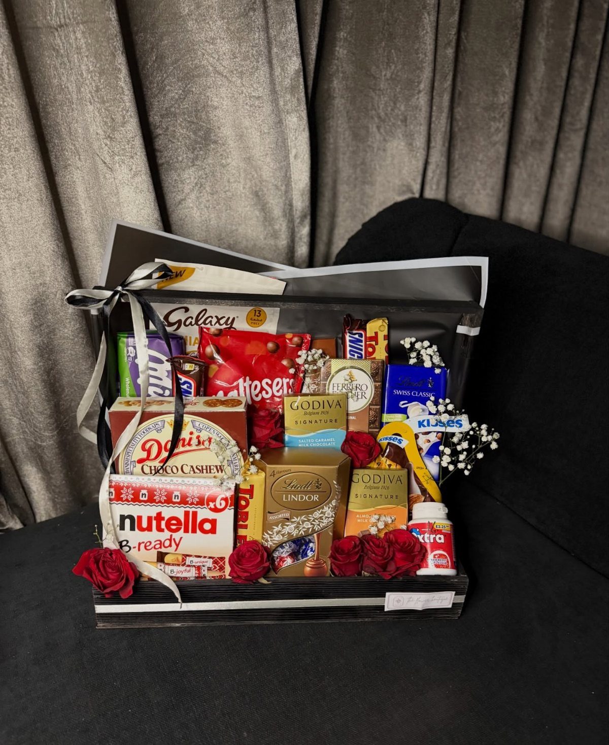 Gift box filled with assorted chocolates, candies, and red roses tied with ribbon.