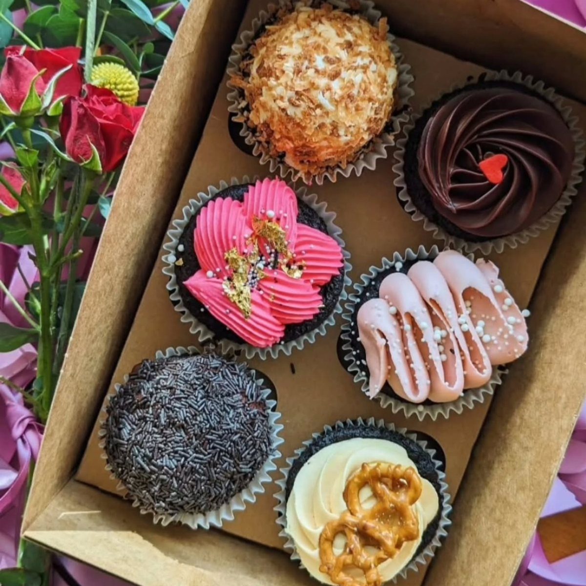 Six decorated chocolate cupcakes in a box with red roses on the side.
