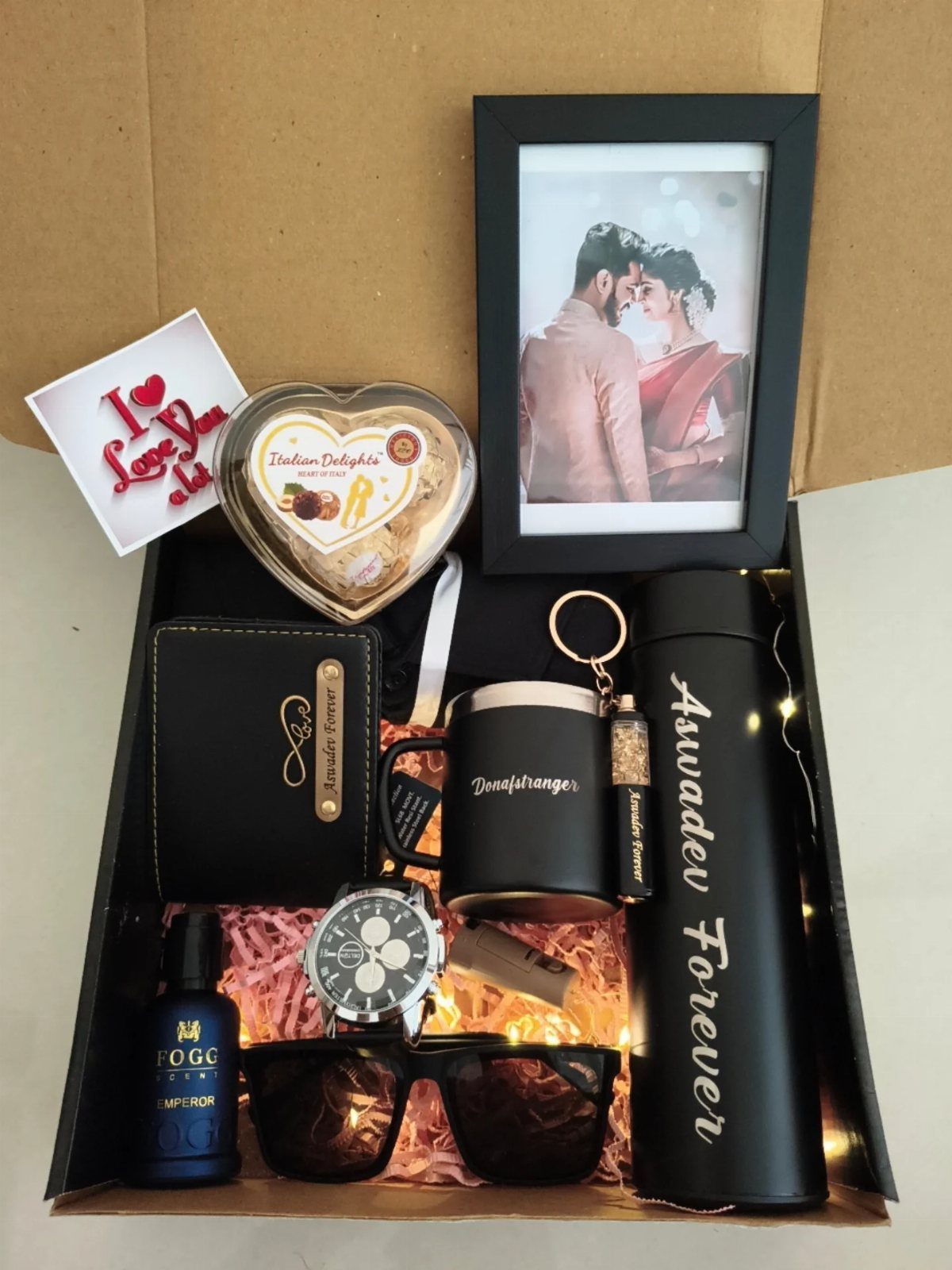Black gift box containing a framed couple's photo, chocolates, watch, and accessories.