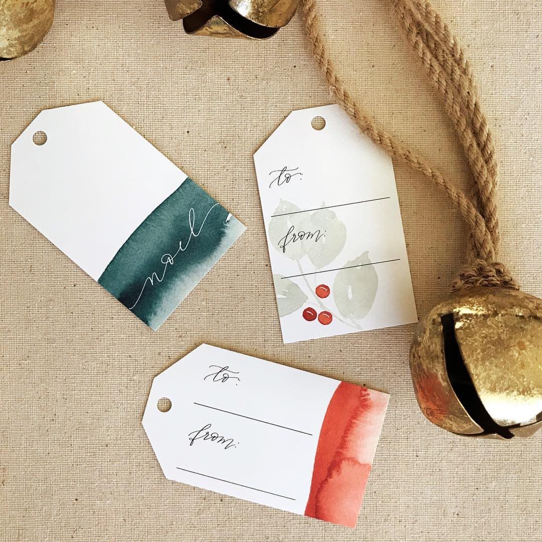 Three watercolor gift tags with festive designs resting on a beige linen fabric.